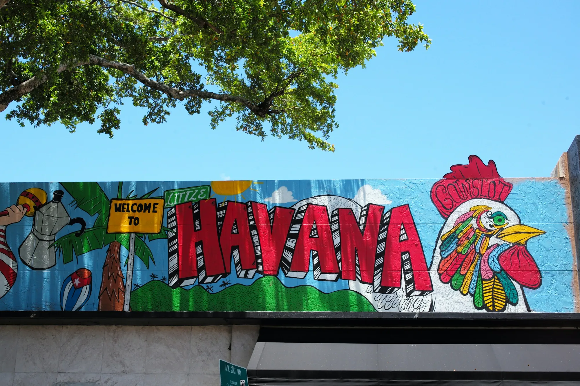 A painted Welcome to Little Havana mural with a rooster, under a shade tree